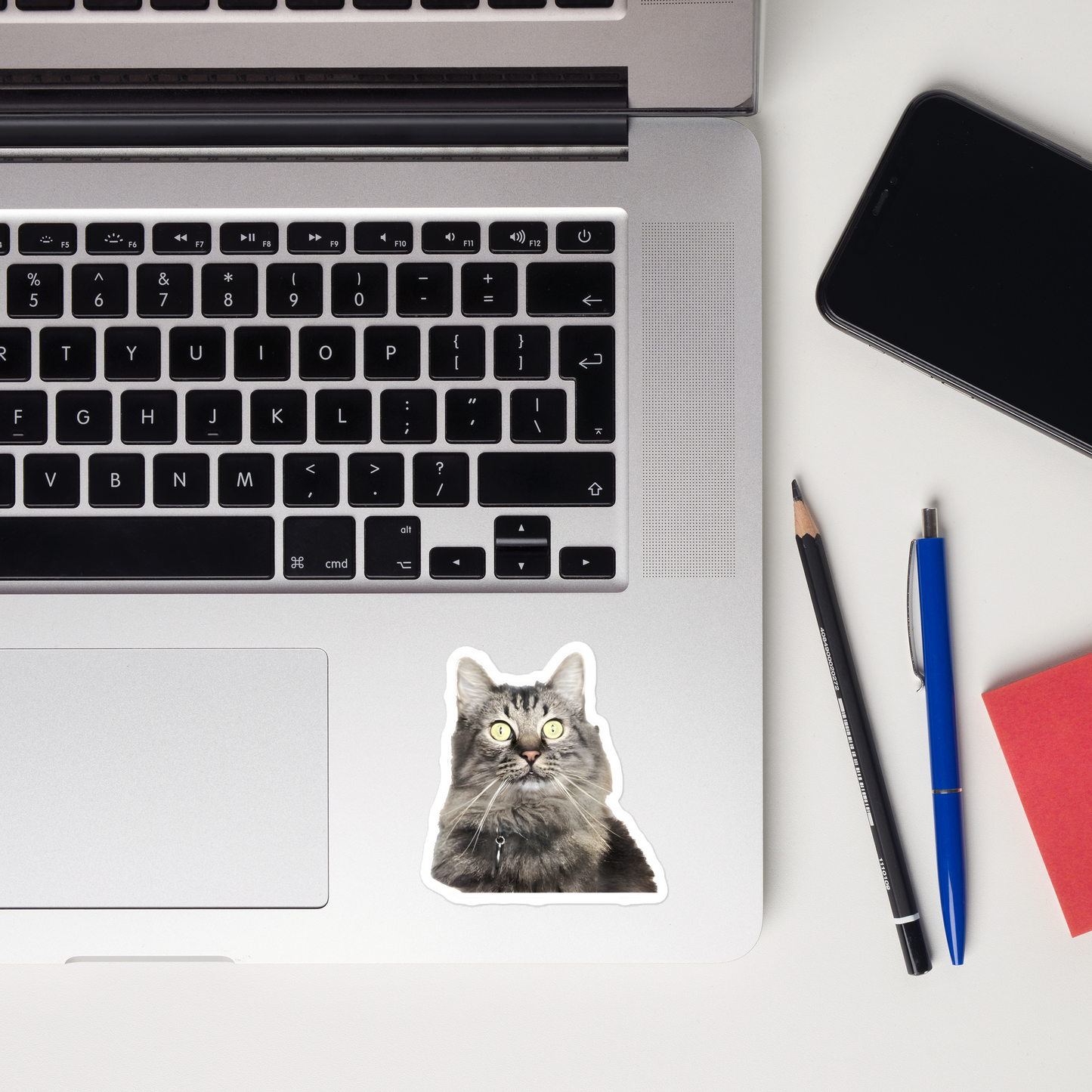 Laptop with a cat sticker, phone, pencil, pen, and notebook on a white surface - 3"x3"