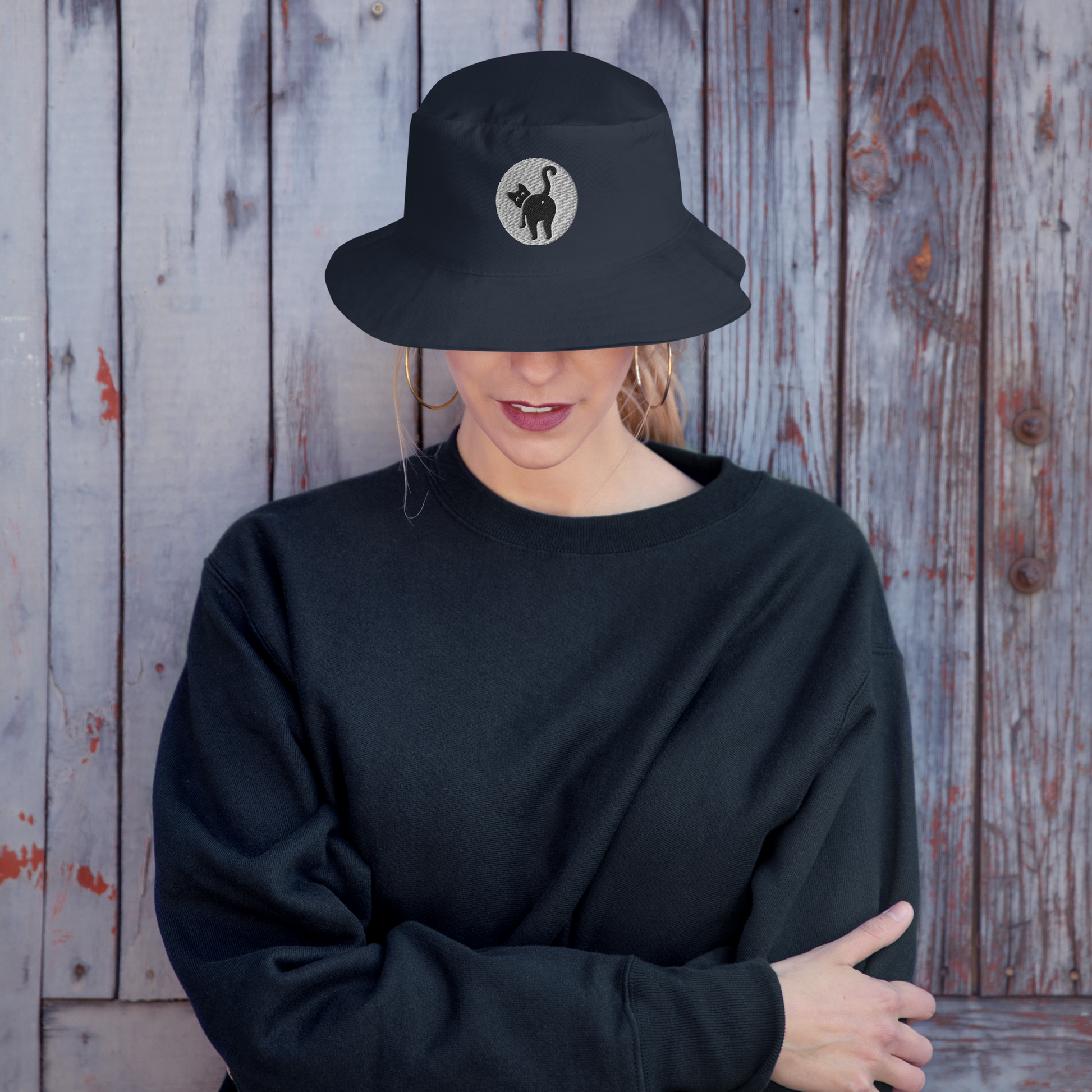 Person wearing a black bucket hat with a logo and a black sweatshirt against a wooden background - navy