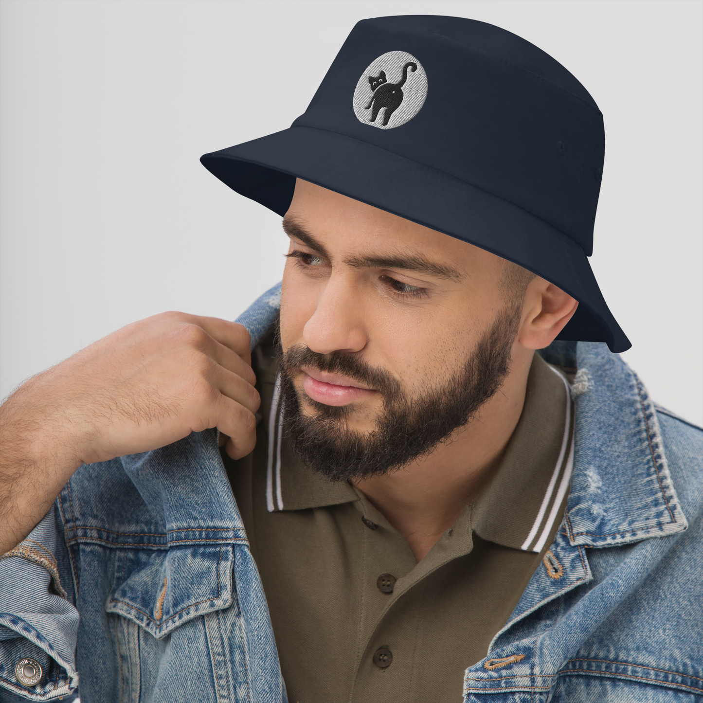 Man wearing a navy bucket hat with a logo, brown polo shirt, and denim jacket on a light gray background - navy