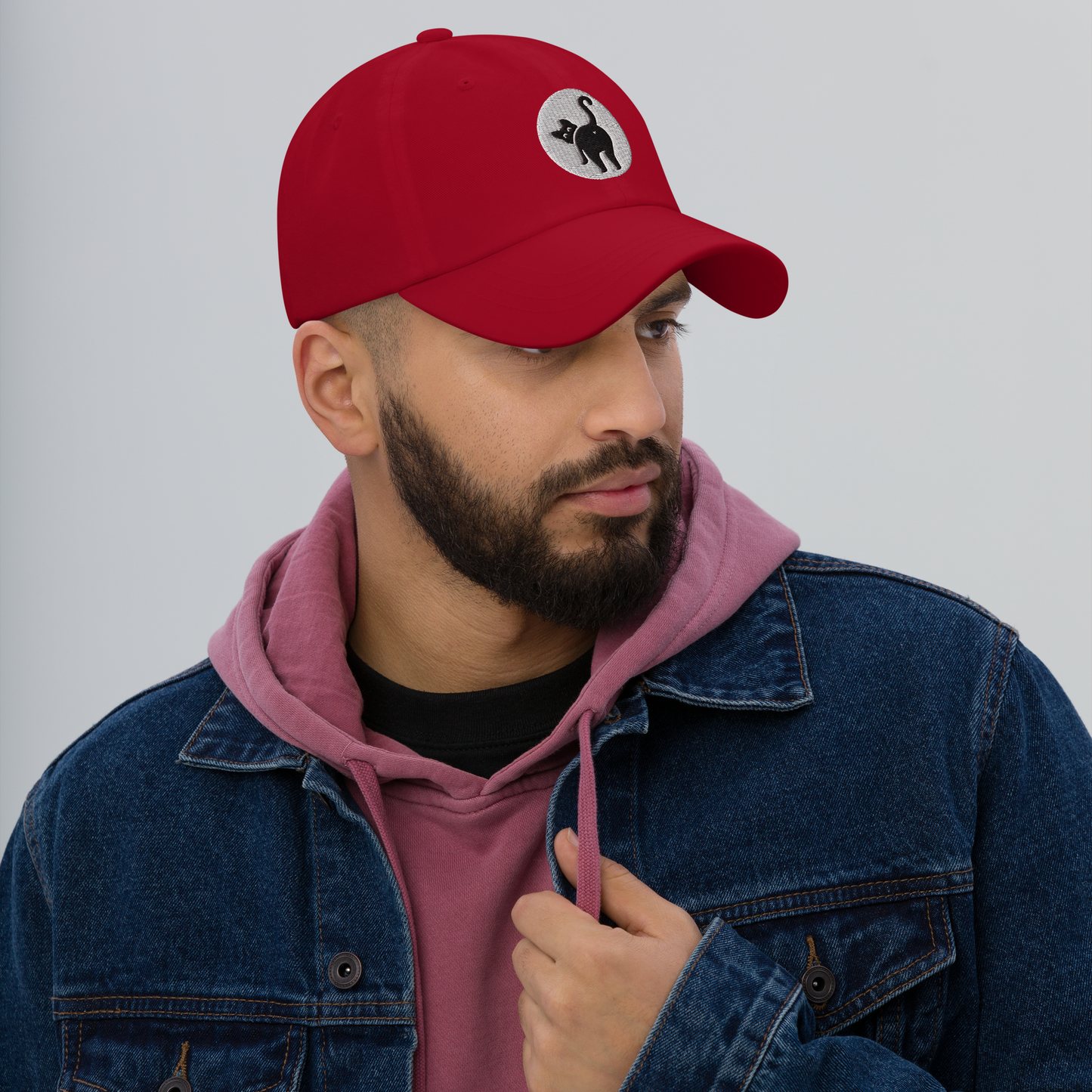 Man wearing a red cap with a black and white logo, denim jacket, and pink hoodie against a gray background - cranberry