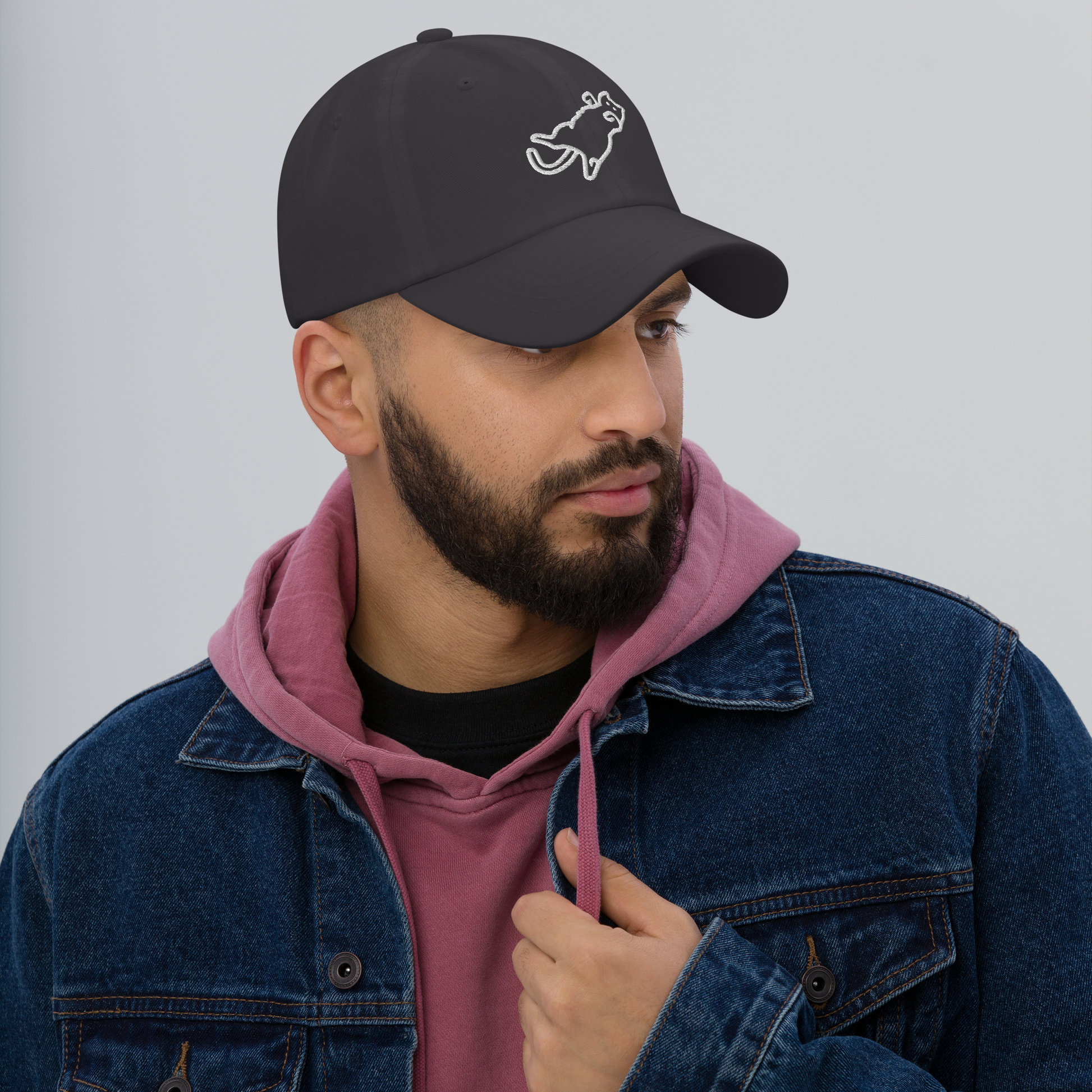Man wearing a black cap with a logo, pink hoodie, and blue denim jacket on a gray background - dark grey