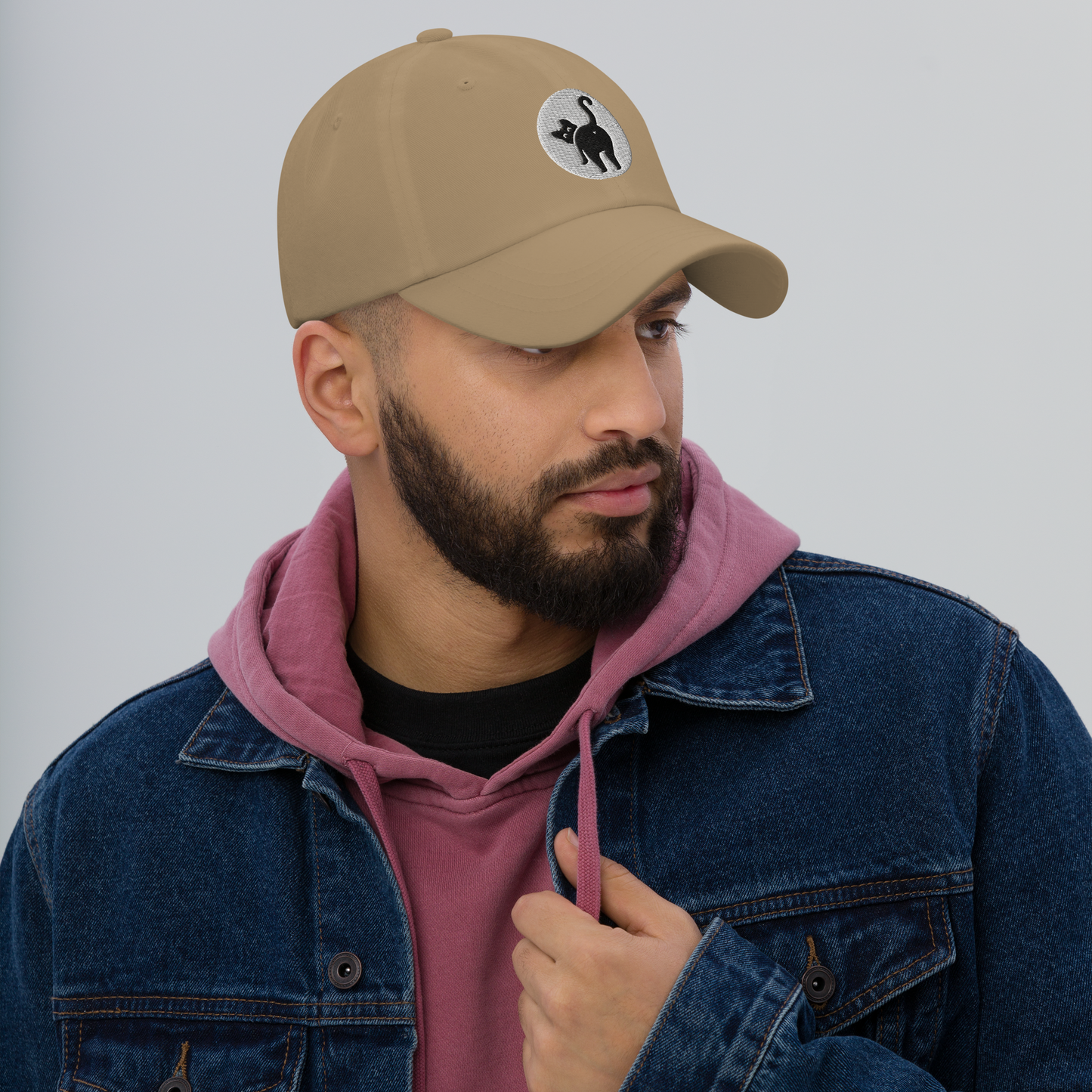 Man wearing a beige cap with a logo, denim jacket, and pink hoodie against a gray background - khaki