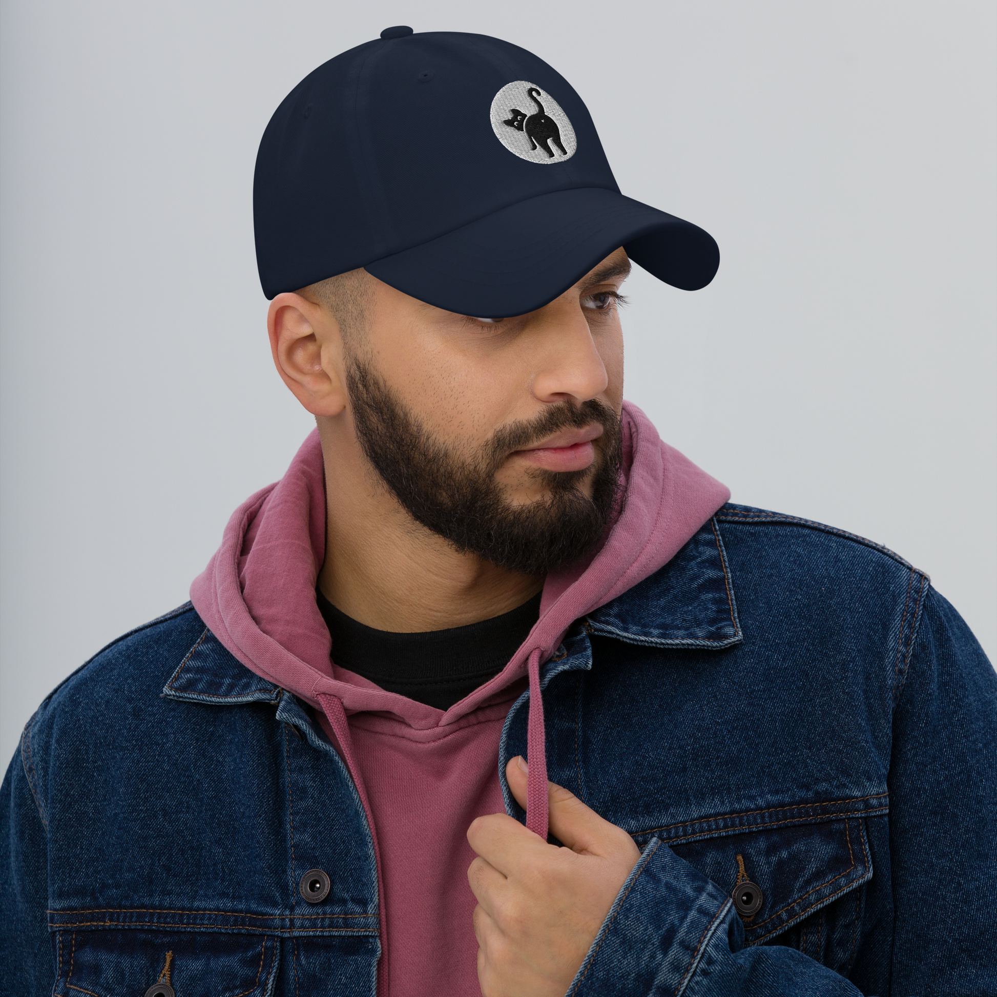 Man wearing a navy cap with a logo, pink hoodie, and denim jacket on a gray background - navy