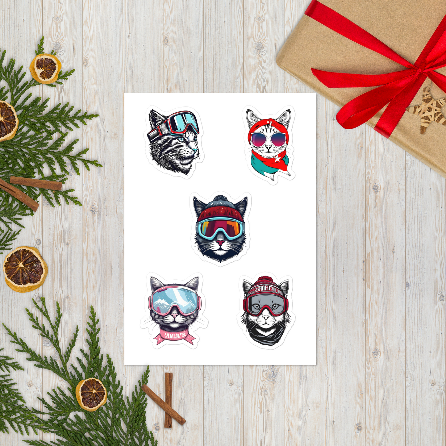 Sheet of temporary tattoos with cat designs wearing colorful goggles on a wooden surface with Christmas decorations.