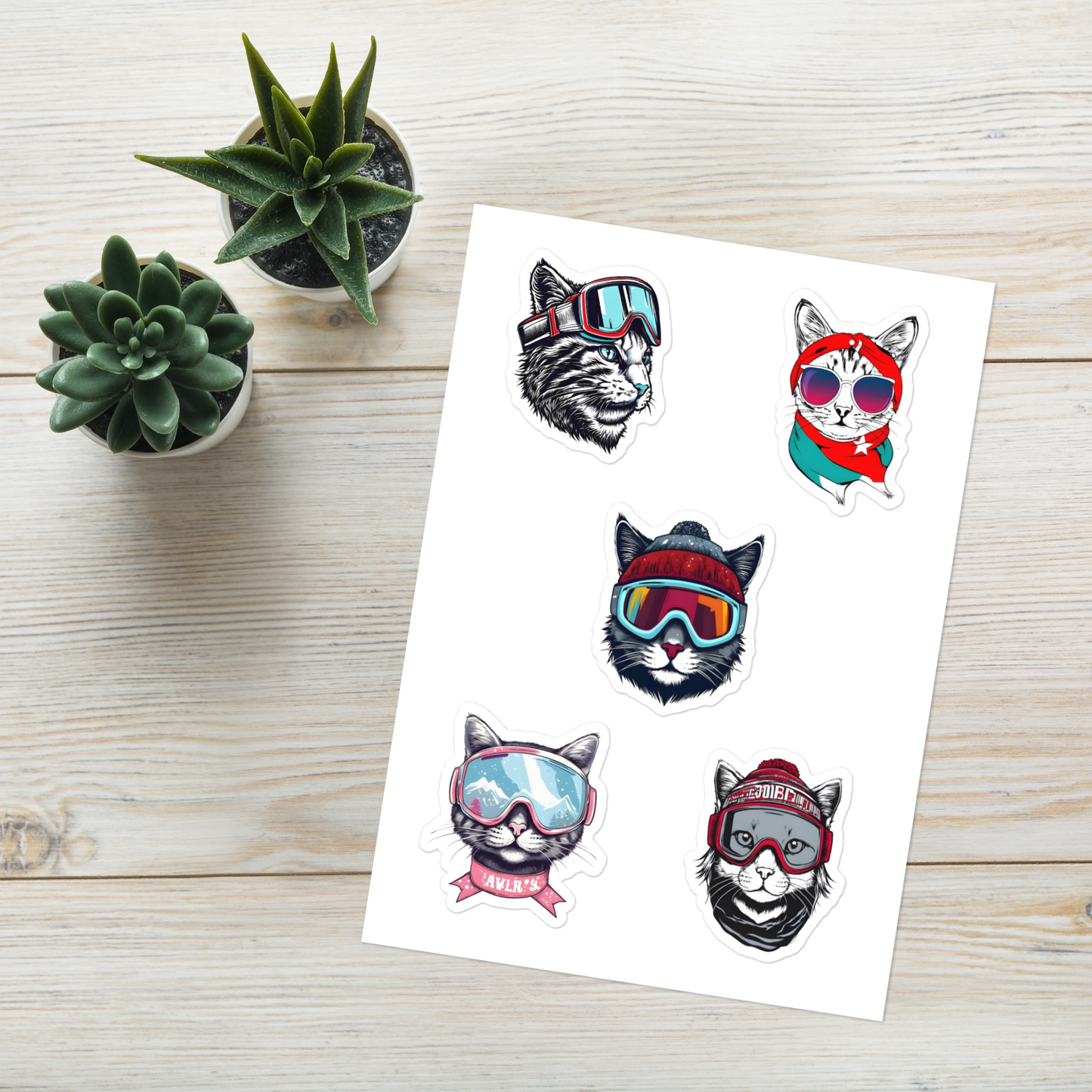 Sheet of cat-themed temporary tattoos with goggles on a wooden surface with small plants.