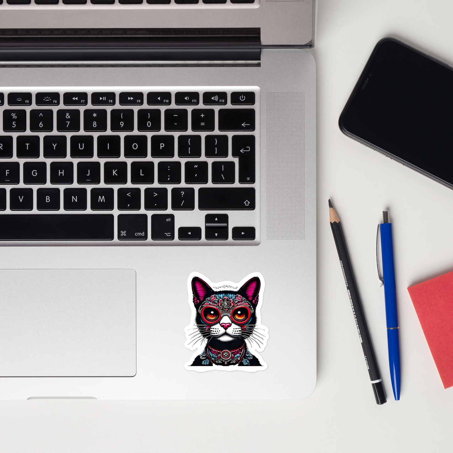 Laptop with a colorful cat sticker on a desk with a phone, pencils, and a notebook. - 3"x3"