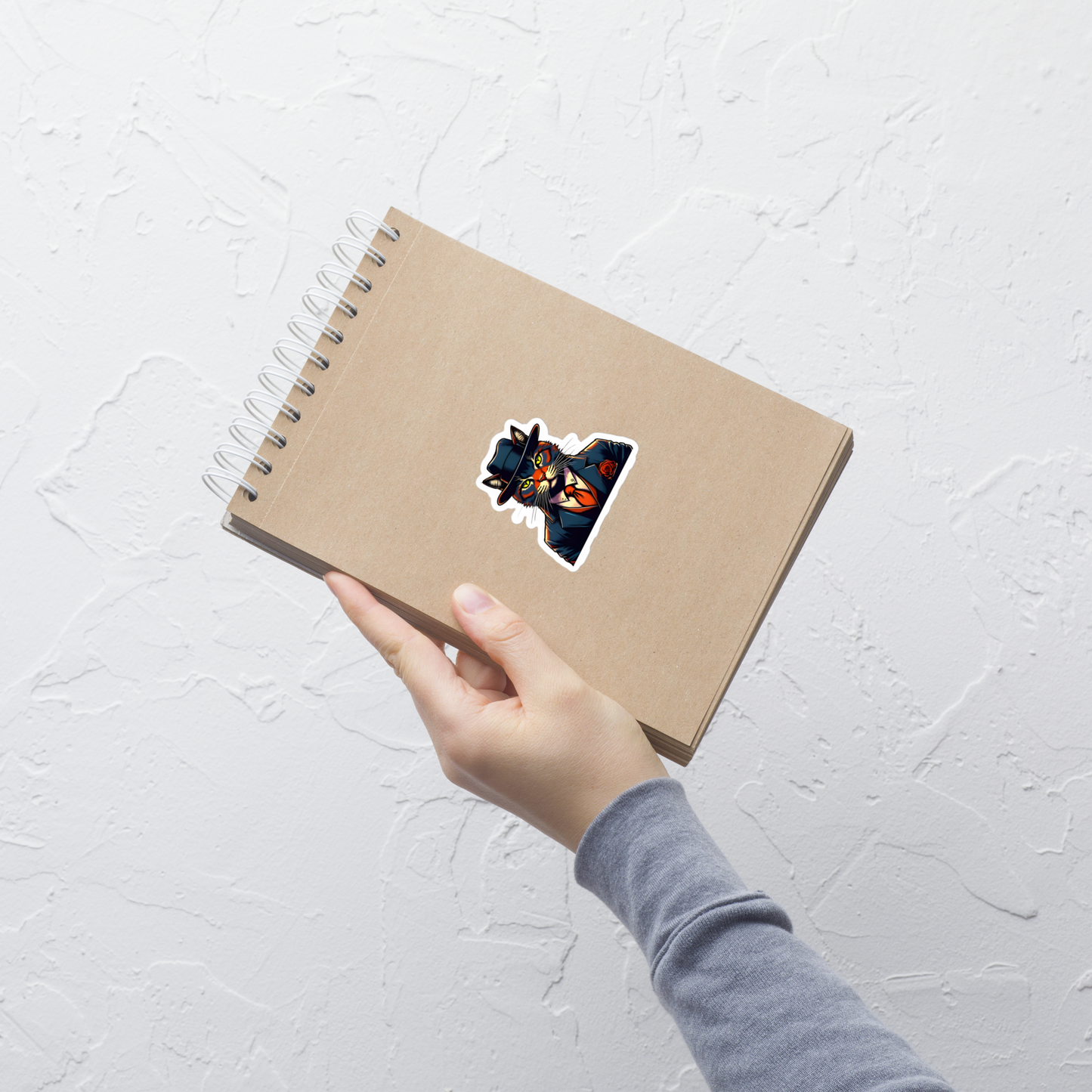 Hand holding a brown notebook with a colorful sticker on a light gray textured background - 3"x3"