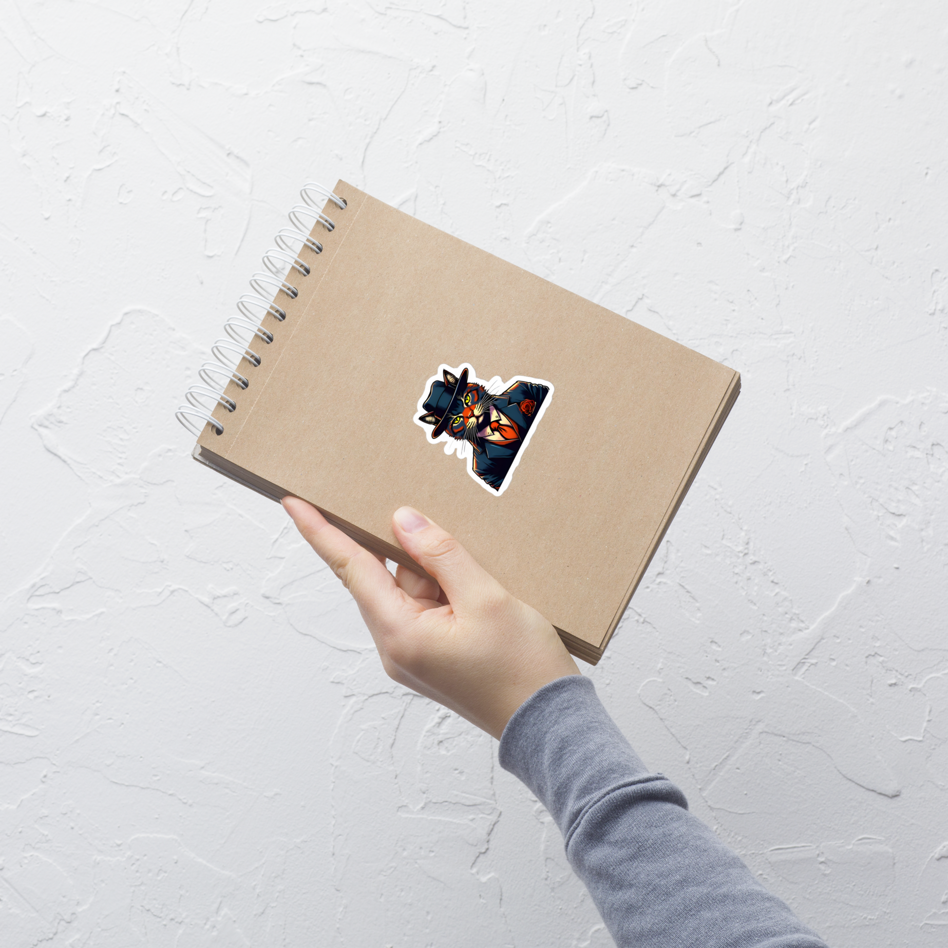 Hand holding a brown notebook with a colorful sticker on a light gray textured background - 3"x3"